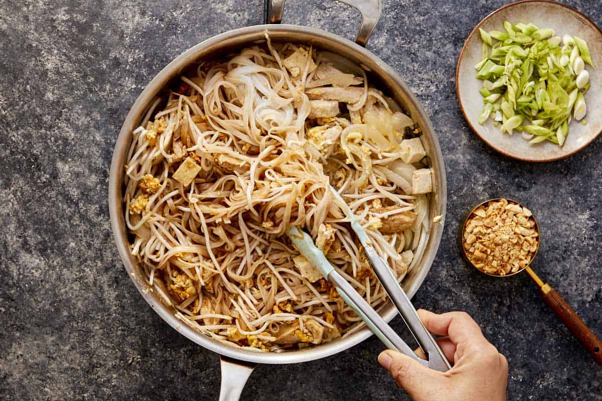 Cooked rice noodles added to the pan with chicken, tofu, eggs, and bean sprouts, tossed with tongs.