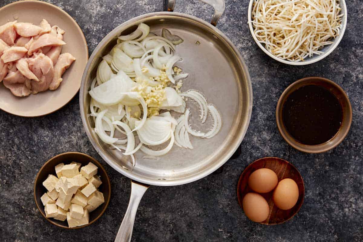 Sliced onion and minced garlic in a pan, with chicken, eggs, tofu, bean sprouts, and sauce on the side