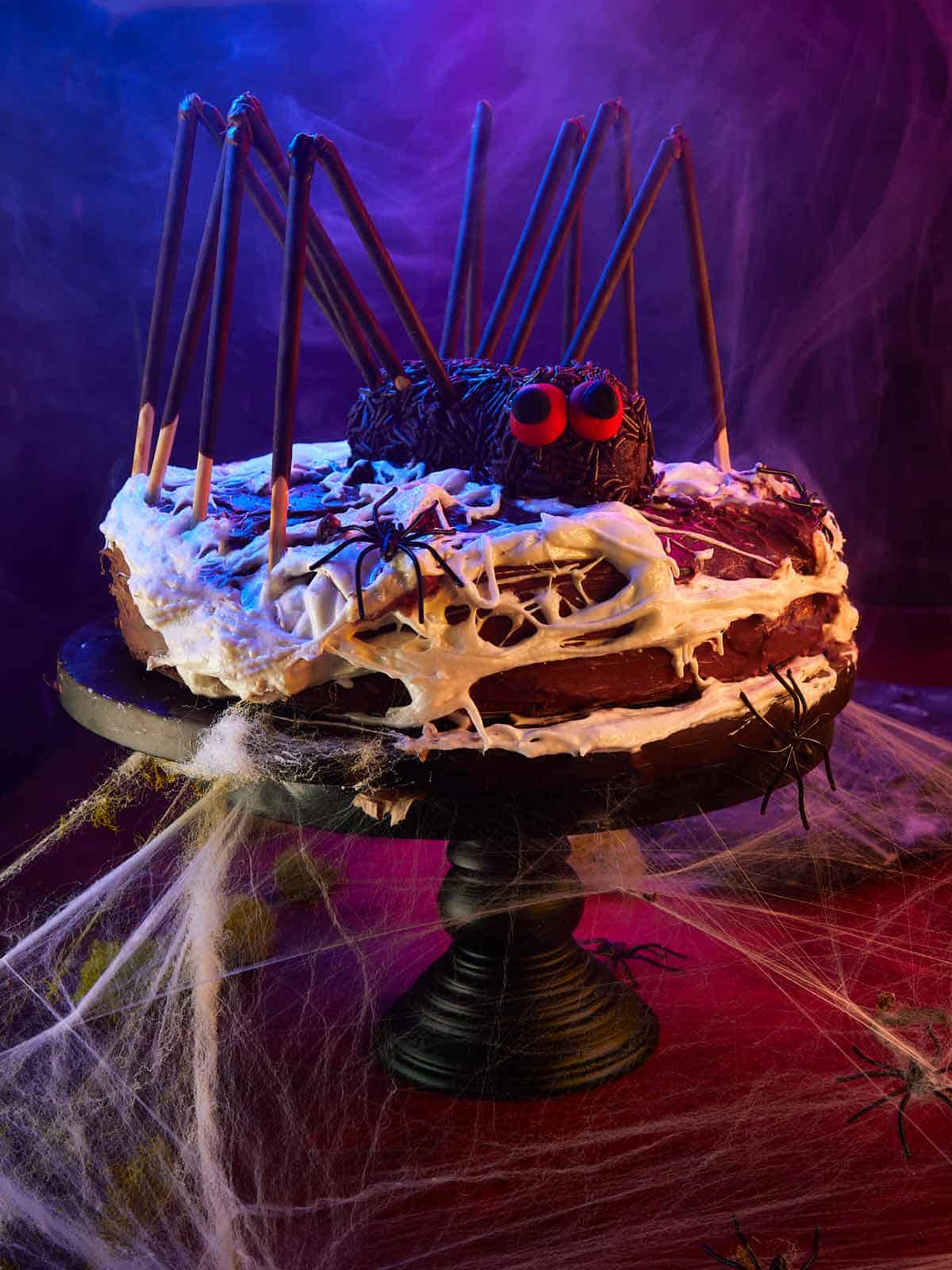 Chocolate Spider Layer Cake on a black cake stand with dramatic lighting, smoke, and glowing purple and blue hues for Halloween.