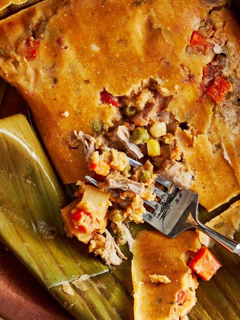 Top-down view of a turquoise Dutch oven filled with stacked tamales wrapped in banana leaves with a fork grabbing some corn