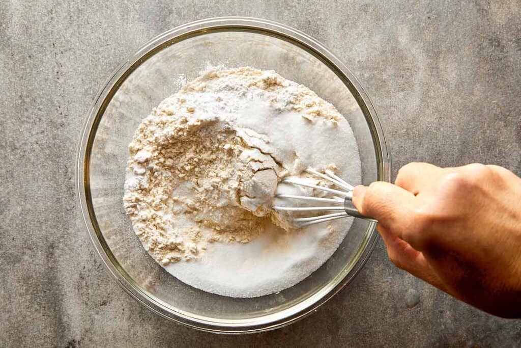A top down shot of a gold whisk, mixing ingredients in a bowl on a counter top with dry ingredients on the bottom of the bowl.