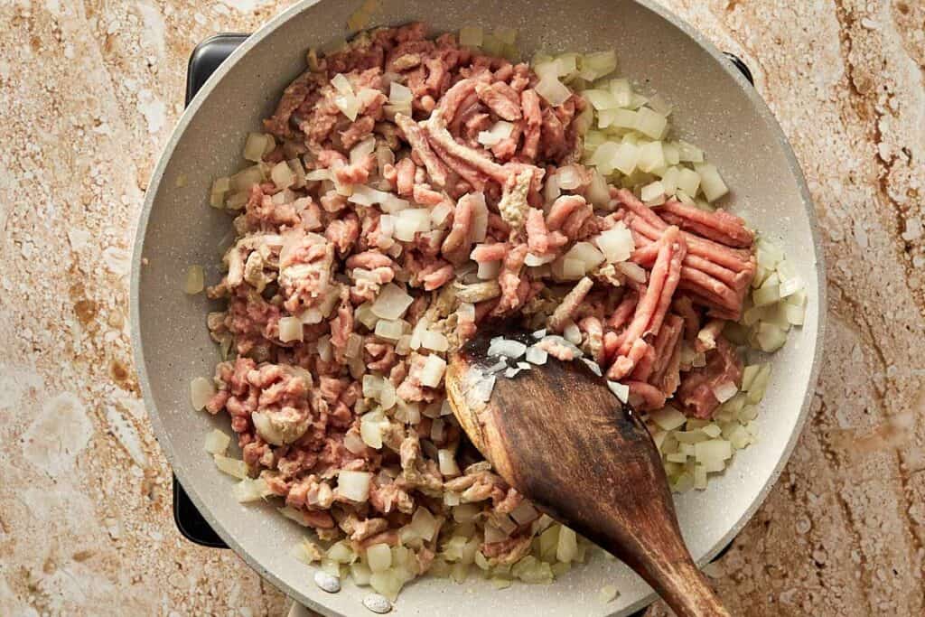 a top down shot of a white pan on a table top with a wooden spoon mixing mince turkey and diced onion, its being fried