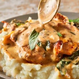 A side view of the tuscan Chicken on a bed of mashed potatoes on top of a green plate with a gold spoon pouring the sauce on top.
