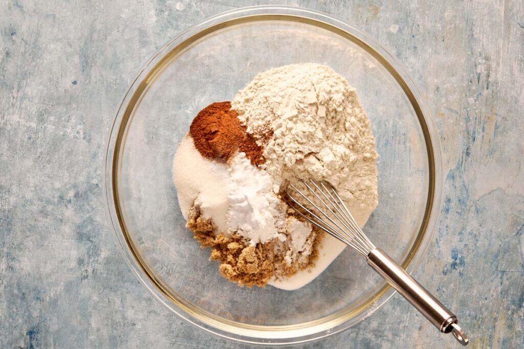 a top down view of a clear glass bowl filled with dry ingredients like flour and cocoa powder with a metal whisk resting on the side of the bowl.