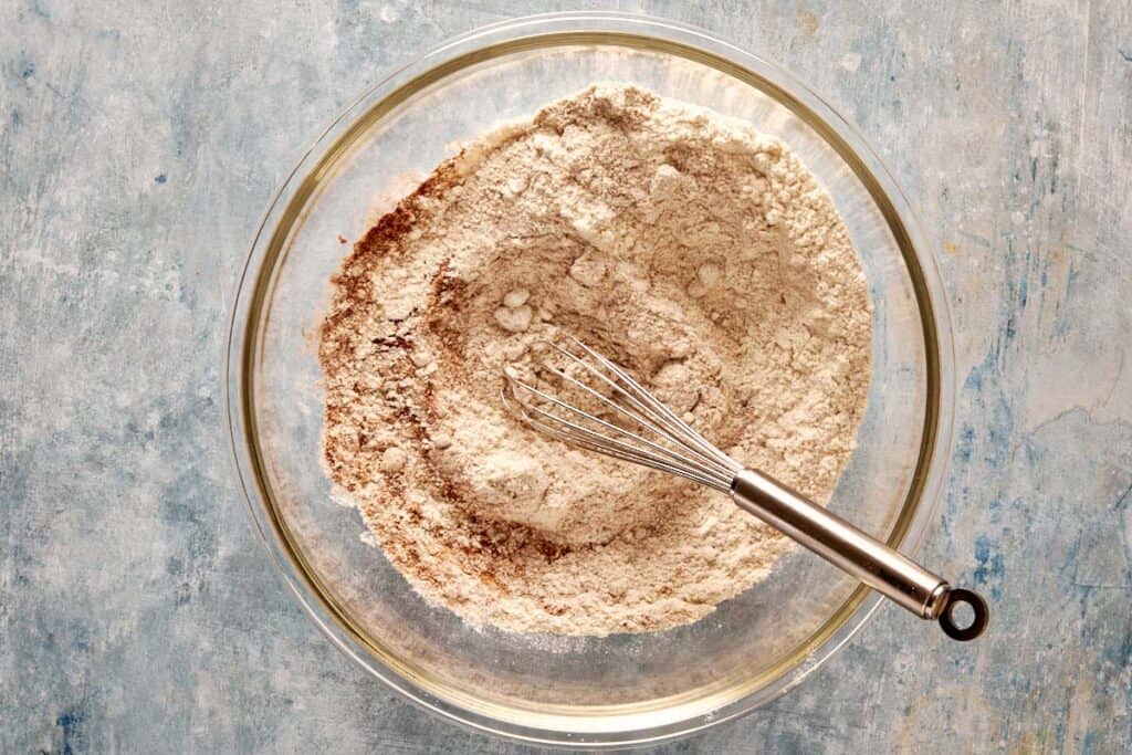 a top down view of a glass bowl with all the dry ingredients mixed with a metal whisk which is currently resting on the side of the bowl.
