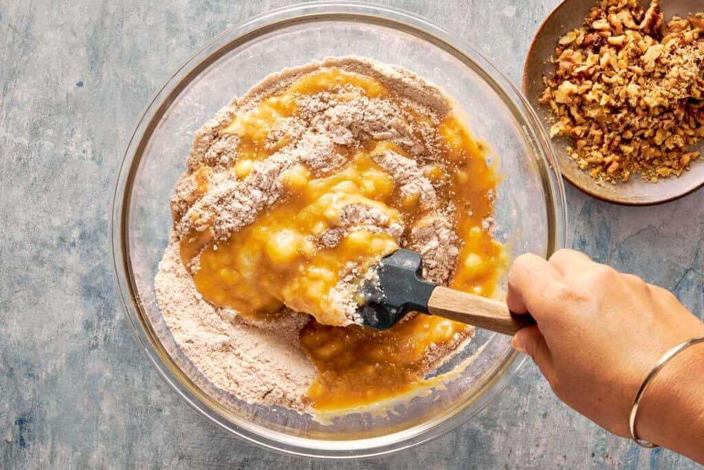 a top down view of a green spatula mixing both the wet ingredients and dry ingredients in a glass bowl sitting on top of a bench