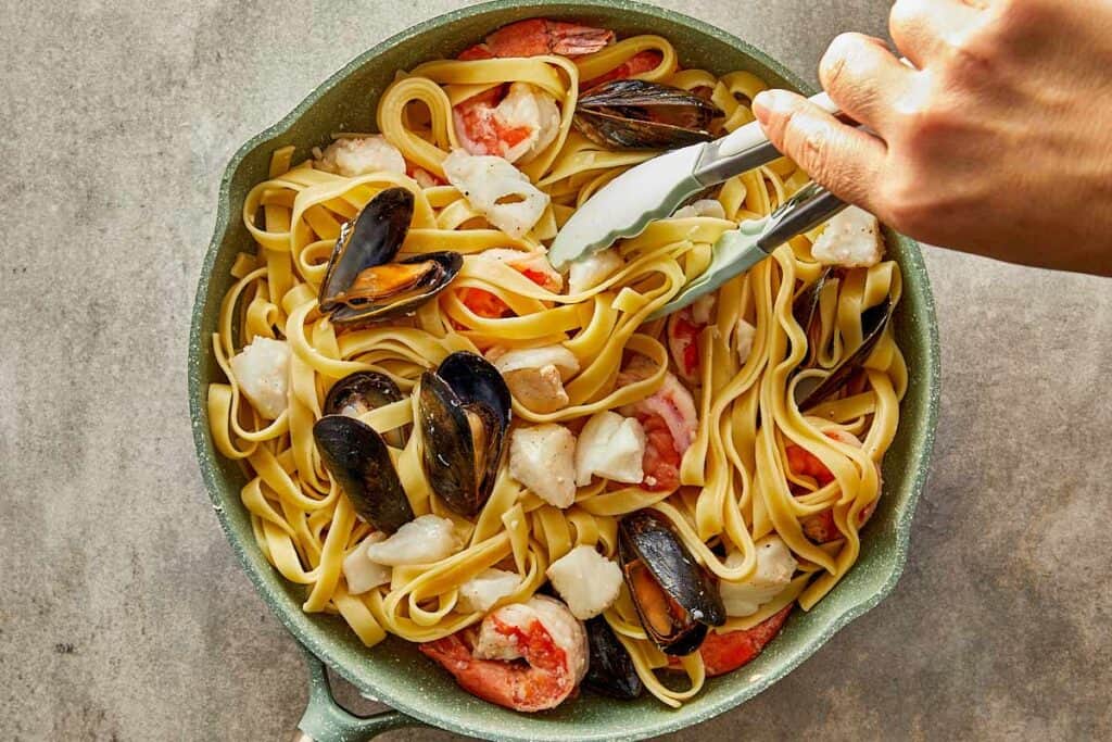 a top down shot of a hand holding tongs picking up pasta and seafood