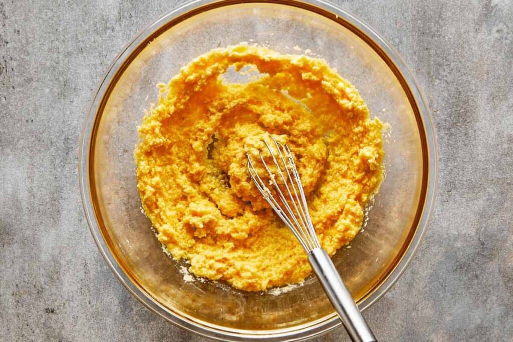 a top down view of a metal whisk in a glass bowl sitting on a grey bench mixing a thick yellow paste which is lumpy