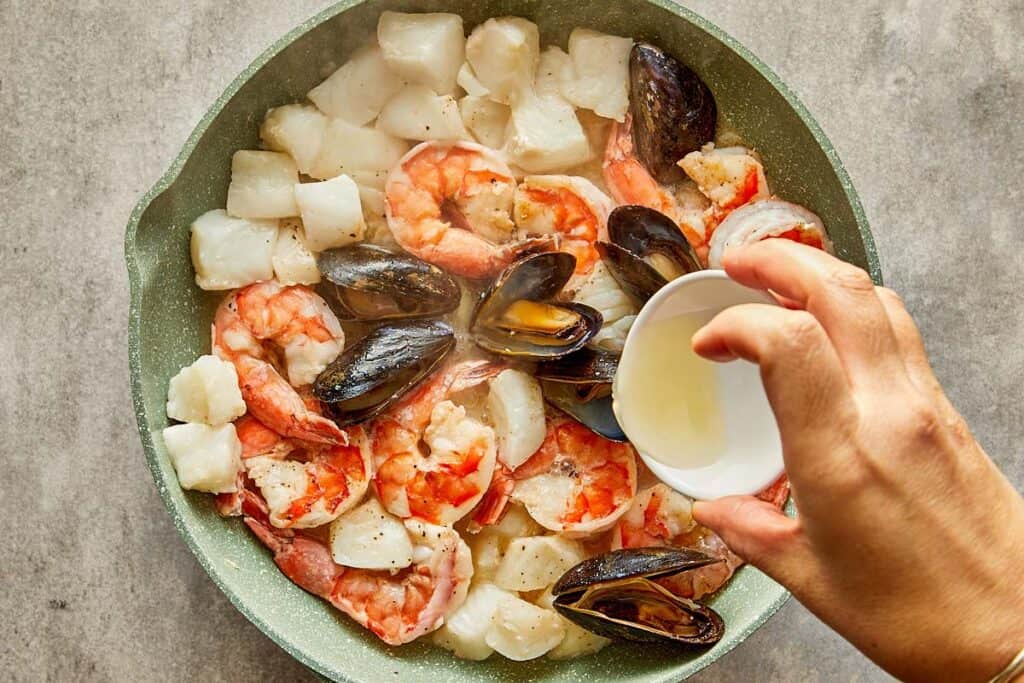 a top down view of seafood in a pan being cooked with what looks like white wine being poured into the pan.
