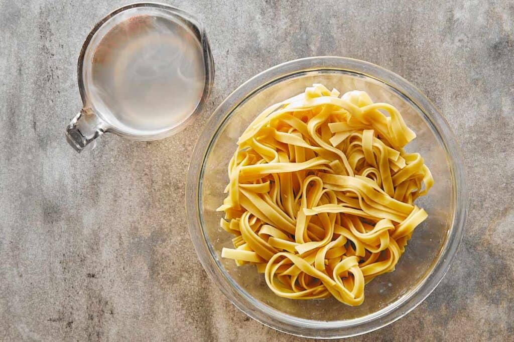 a top down view of a glass bowl full of cooked pasta and a jug of water next to it.
