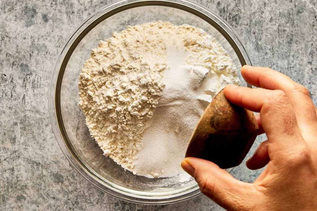 A top down view of a glass bowl on a kitchen bench filled with flour and a cup of sugar being poured into it by a female hand