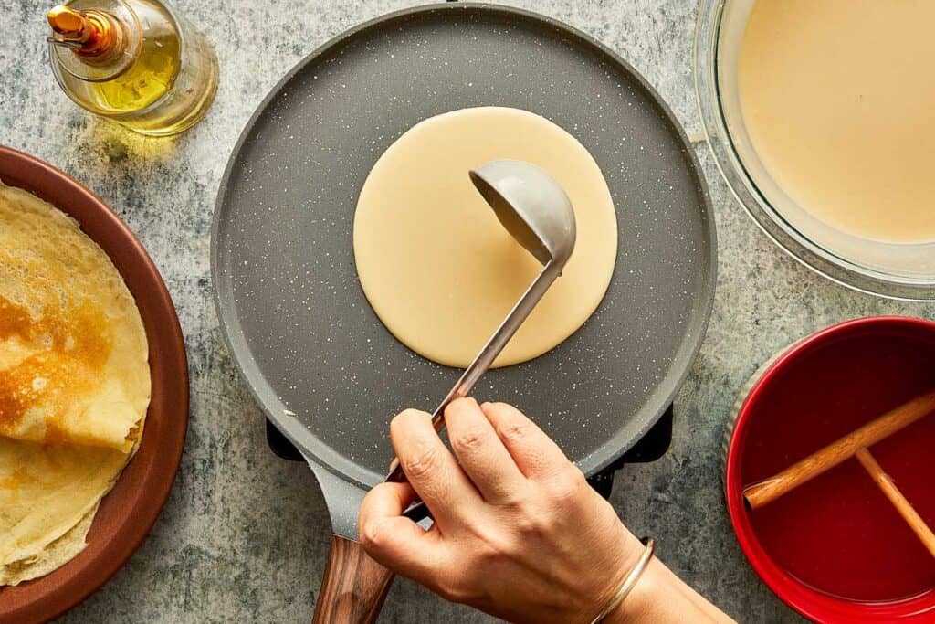 a top down view of a ladle of batter being poured into a crepe pan