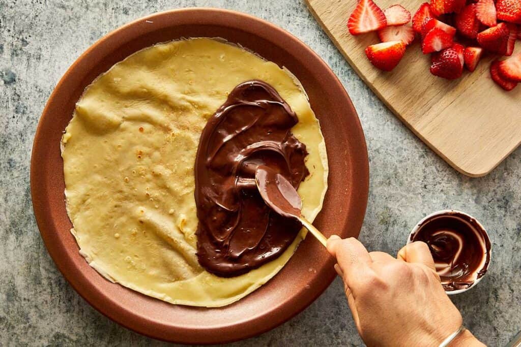 a cooked crepe sitting on top of a ceramic brown plate, the crepe is creased as it sits on the plate and a golden spoon is covering one side of the crepe with Nutella paste