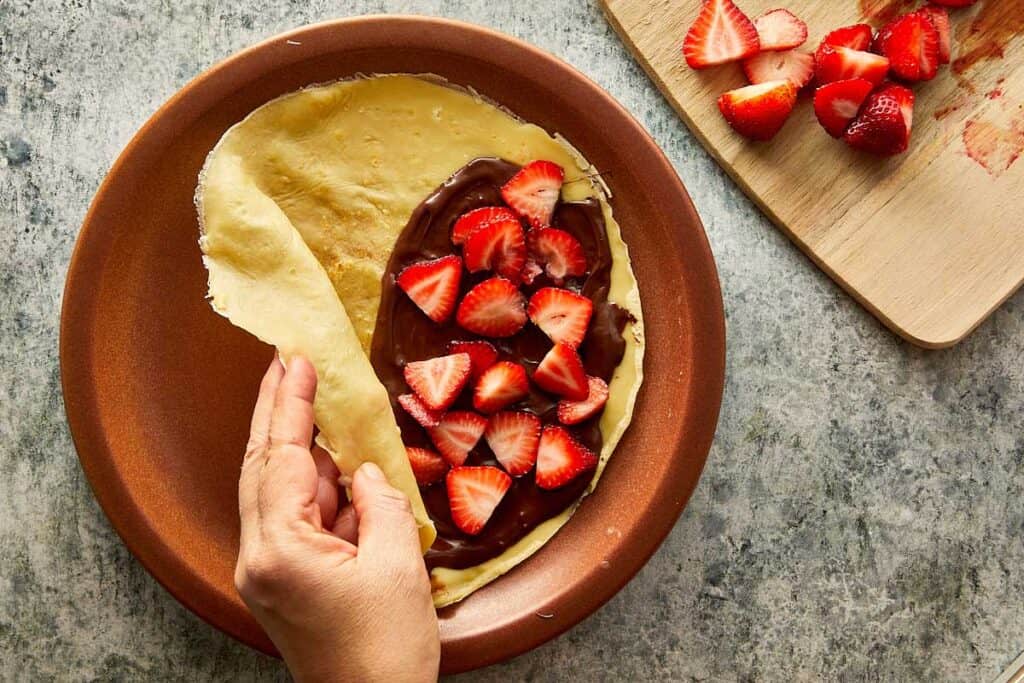 a cooked crepe sitting on top of a ceramic brown plate, the crepe is creased as it sits on the plate with nutella paste on one side covered with strawberries on top which have been quartered