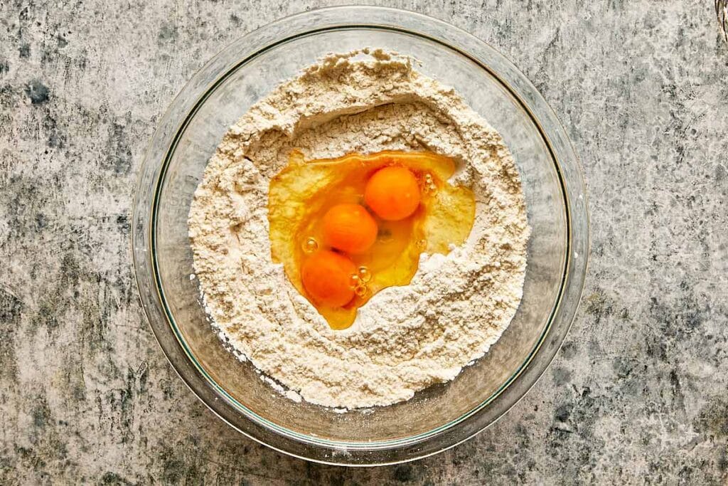 a top down view of a glass mixing bowl on a kitchen bench with 3 eggs in the middle of a flour mix