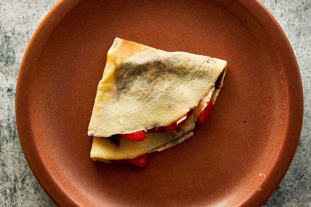 a top down view of a brown ceramic plate with a folded crepe filled with nutella and strawberries.