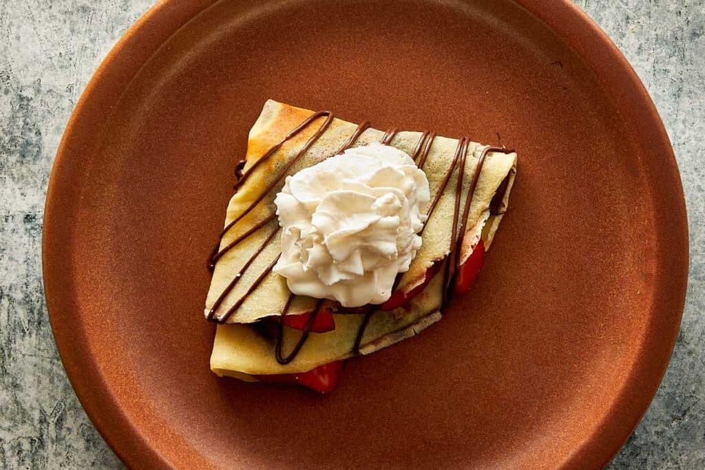 a top down view of a ceramic brown plate with a quartered crepe sitting on top with a blob of cream and some drizzle zig zagged topping of Nutella