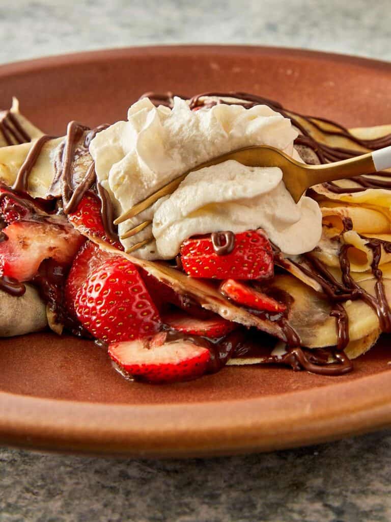 a side view of a ceramic brown plate with a quartered crepe sitting on top with a blob of cream and some drizzle zig zagged topping of Nutella and strawberries on top