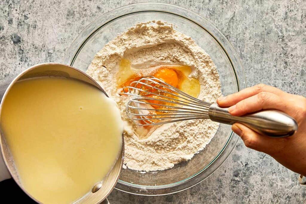 a top down view of a glass mixing bowl on a bench filled with flour and egg mixture and a bowl of milk being poured into it slowly while a hand is whisking everything together.
