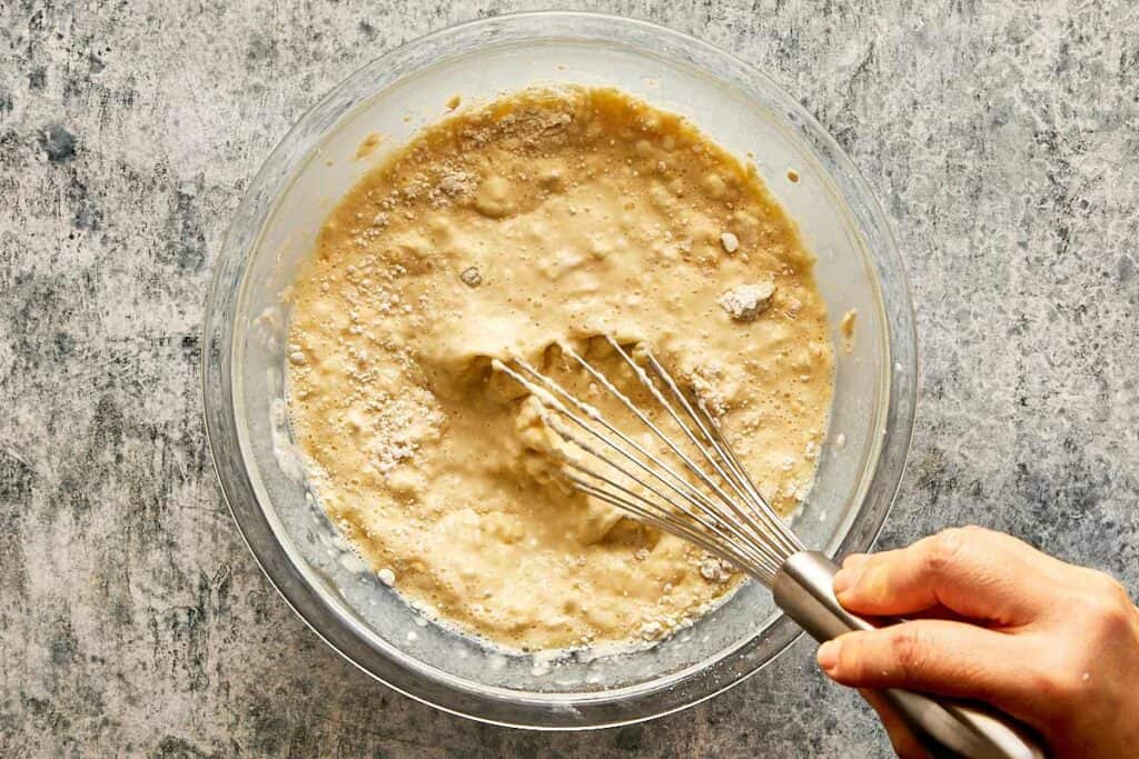 a top down view of a glass bowl and a whisk being held by a female hand mixing a thick white mixture which is lumpy