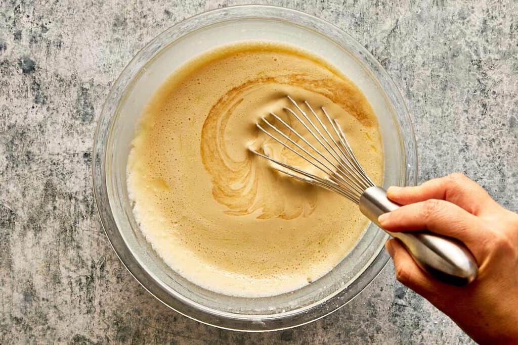 a top down view of a glass bowl and a whisk being held by a female hand mixing a thick white mixture which is getting smooth