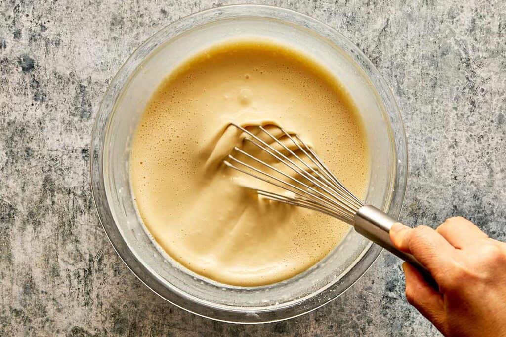 a top down view of a glass bowl and a whisk being held by a female hand mixing a thick white mixture which is smooth