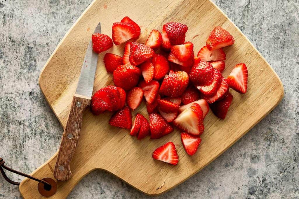 a top down view of a wooden chopping board and strawberries cut into wedges with a small pairing knife