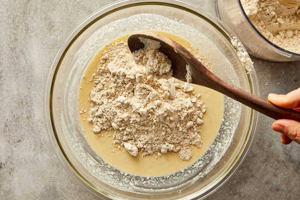 a top down image of a glass mixing bowl with white batter in the bottom of the bowl and white powder on top getting ready to be mixed with a wooden mixing spoon