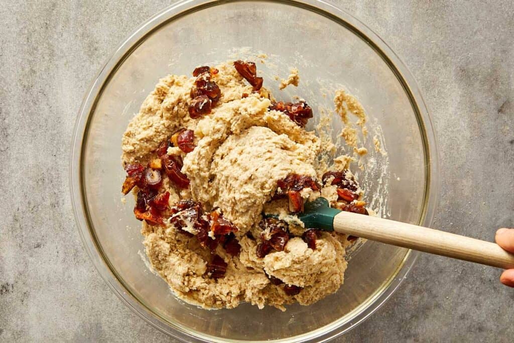 a top down image of a glass mixing bowl with white batter in the bottom of the bowl and white powder on top getting ready to be mixed with a wooden mixing spoon with fruit inside