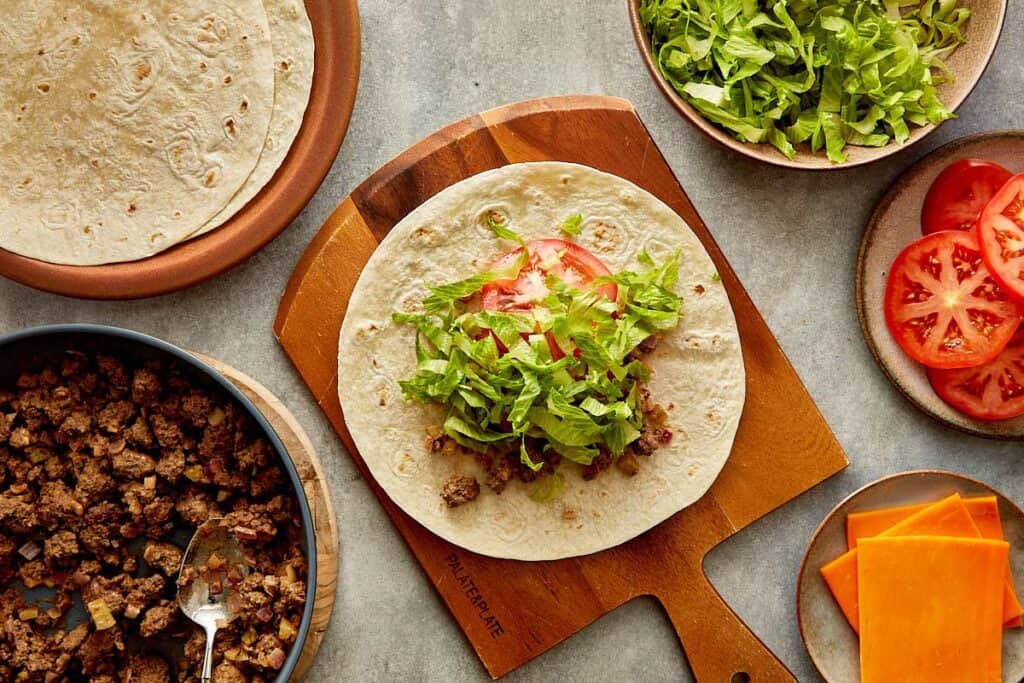 a top down view of a wrap on top of a wooden board sitting on a kitchen bench, the wrap has cooked mince, slice of cheese, lettuce and tomatoes