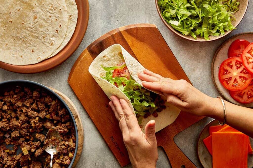 a top down view of a wrap being made by two female hands, the wrap is filled with mince meat and lettuce and its on top of a wooden board.