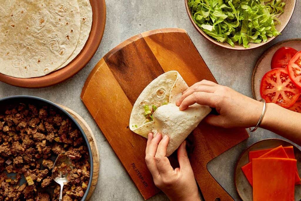 a top down view of a wrap being made by two female hands, the wrap is filled with mince meat and lettuce and its on top of a wooden board.