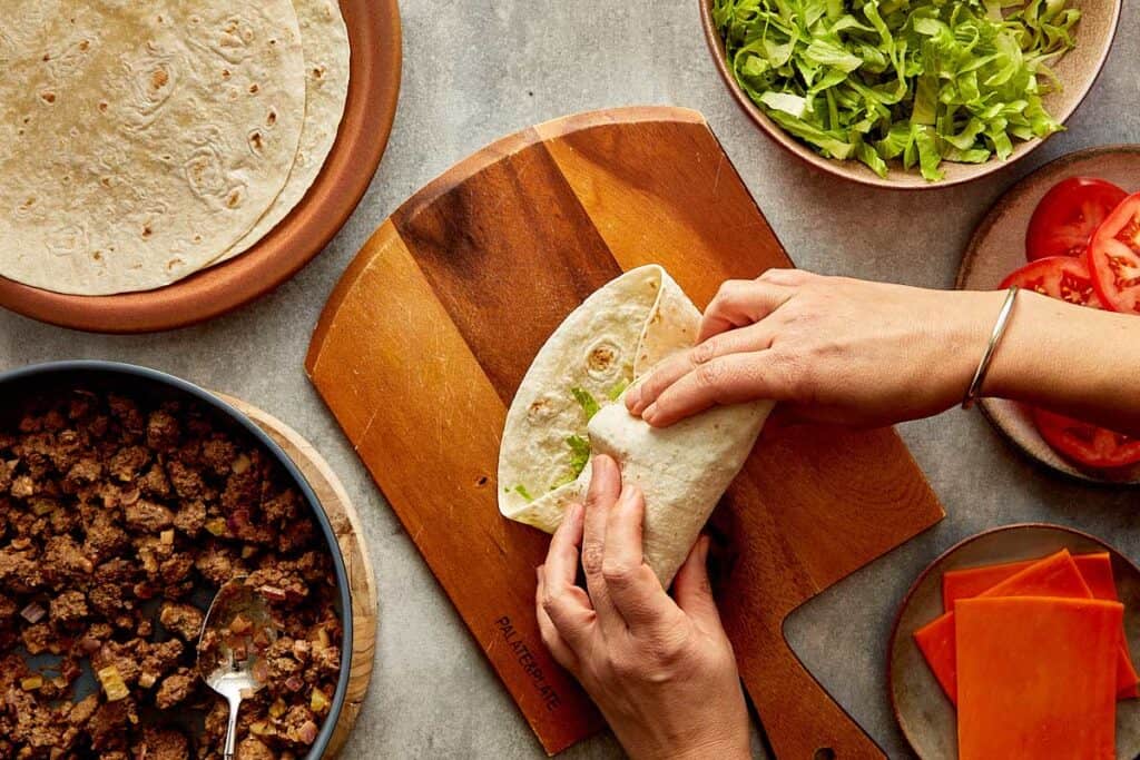 a top down view of a wrap being made by two female hands, the wrap is filled with mince meat and lettuce and its on top of a wooden board.