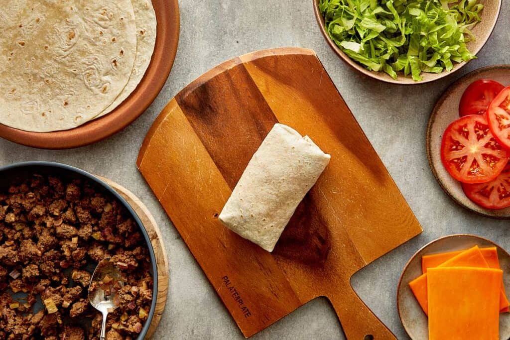 a top down view of a wrap being made by two female hands, the wrap is filled with mince meat and lettuce and its on top of a wooden board.