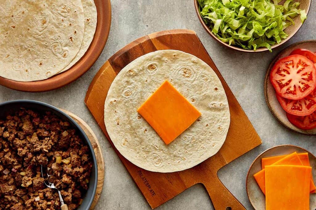 a top down view of a wrap on top of a wooden board sitting on a kitchen bench, the wrap has a slice of cheese on it.