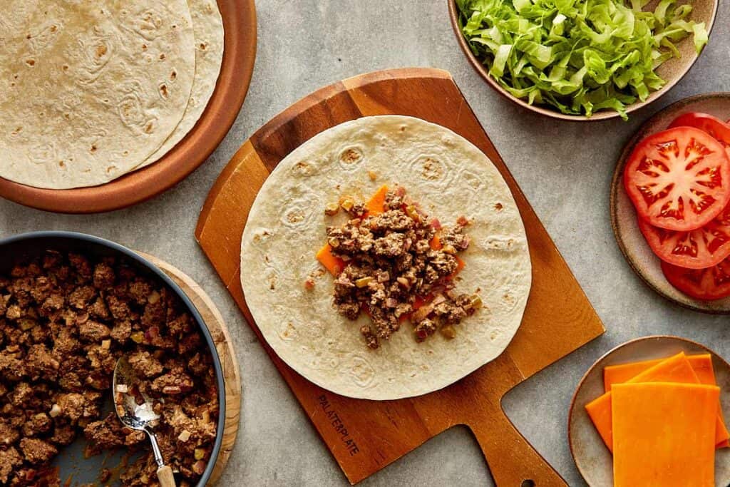 a top down view of a wrap on top of a wooden board sitting on a kitchen bench, the wrap has a slice of cheese and some cooked mince on it