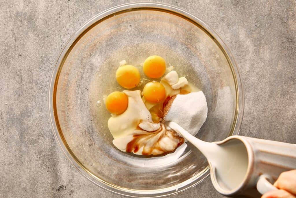 a top down shot showing a glass bowl with eggs and vanilla essence with a jug of milk being poured into it.