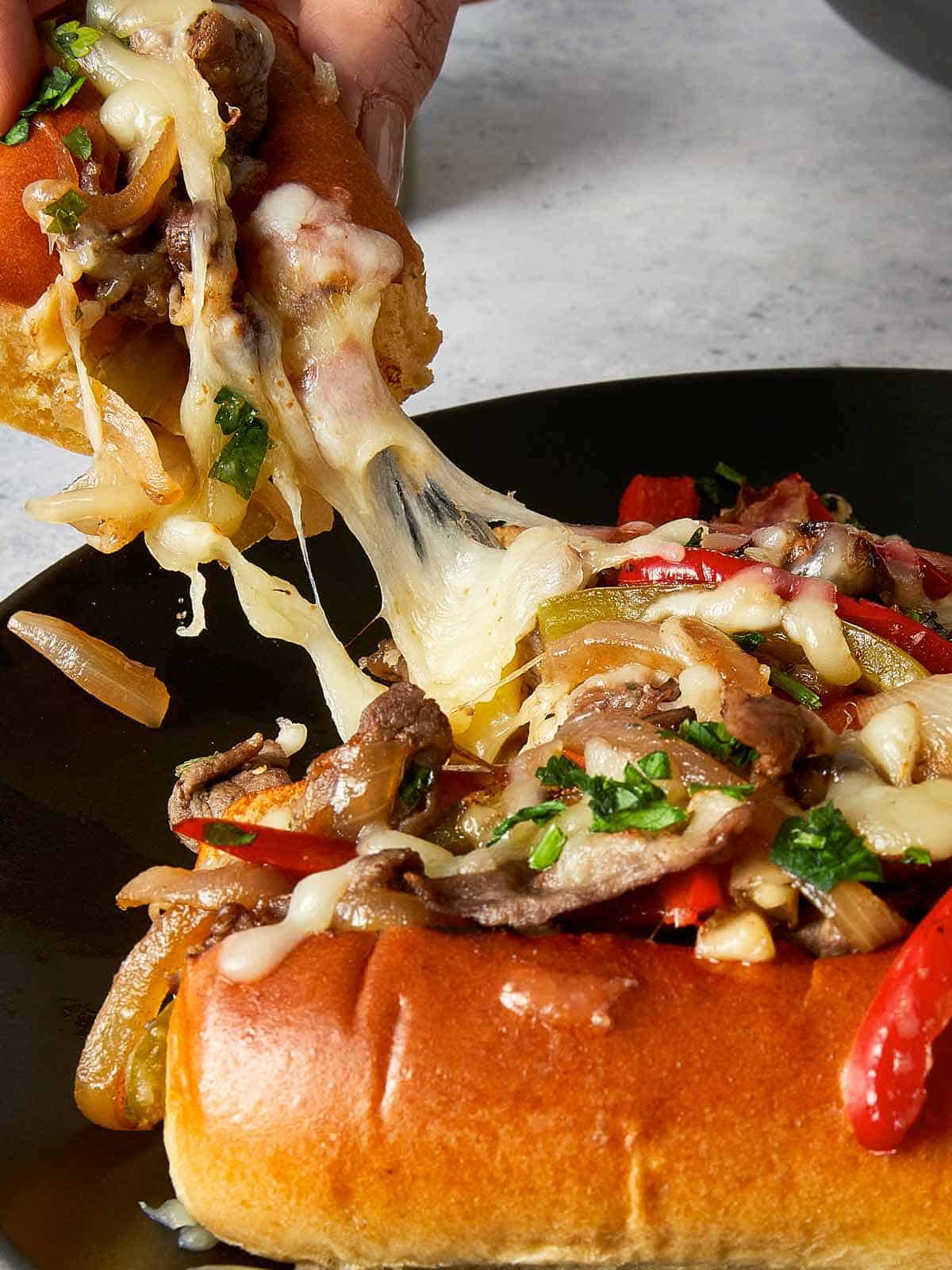 Close up of Philly cheesesteak sandwich with melted mozzarella cheese pull, sliced steak, sautéed onions, bell peppers, and parsley served in a toasted hoagie roll.