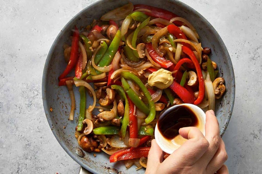 Worcestershire sauce and Dijon mustard being added to sautéed onions, bell peppers, mushrooms, and garlic in the skillet to build flavor.