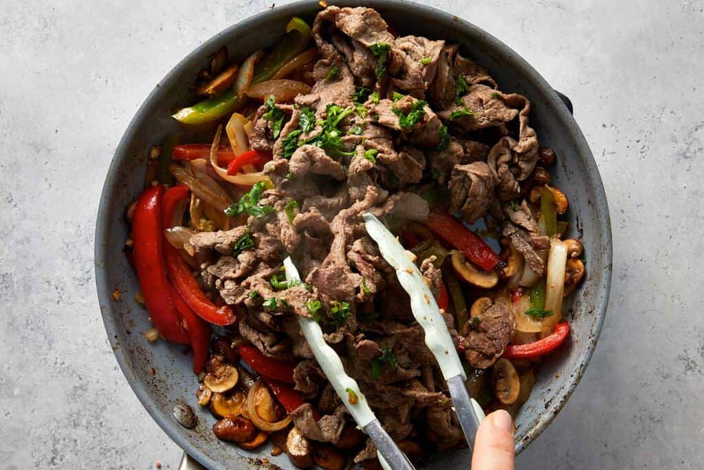 Cooked steak added back into the skillet with sautéed onions, bell peppers, mushrooms, and parsley, being tossed together to combine before adding cheese.