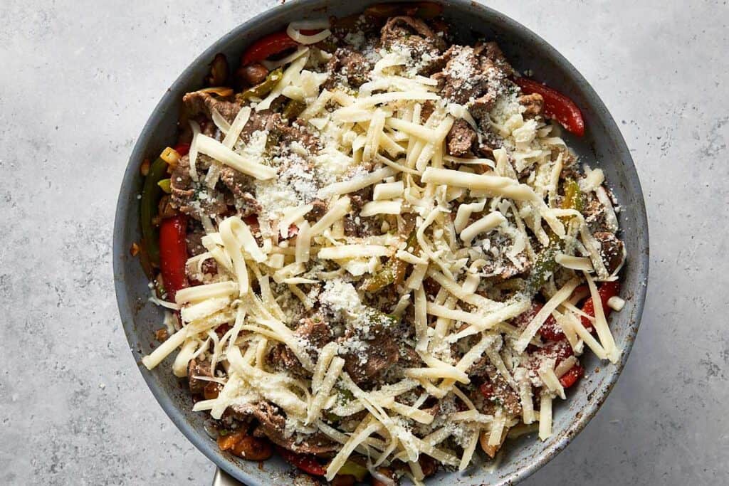 Shredded mozzarella and grated Pecorino sprinkled over the steak, peppers, onions, and mushrooms in the skillet, ready to be covered and melted.