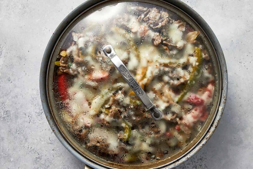 Skillet covered with a glass lid as the steak, peppers, onions, and mushrooms steam while the cheese melts until bubbly and fully melted.
