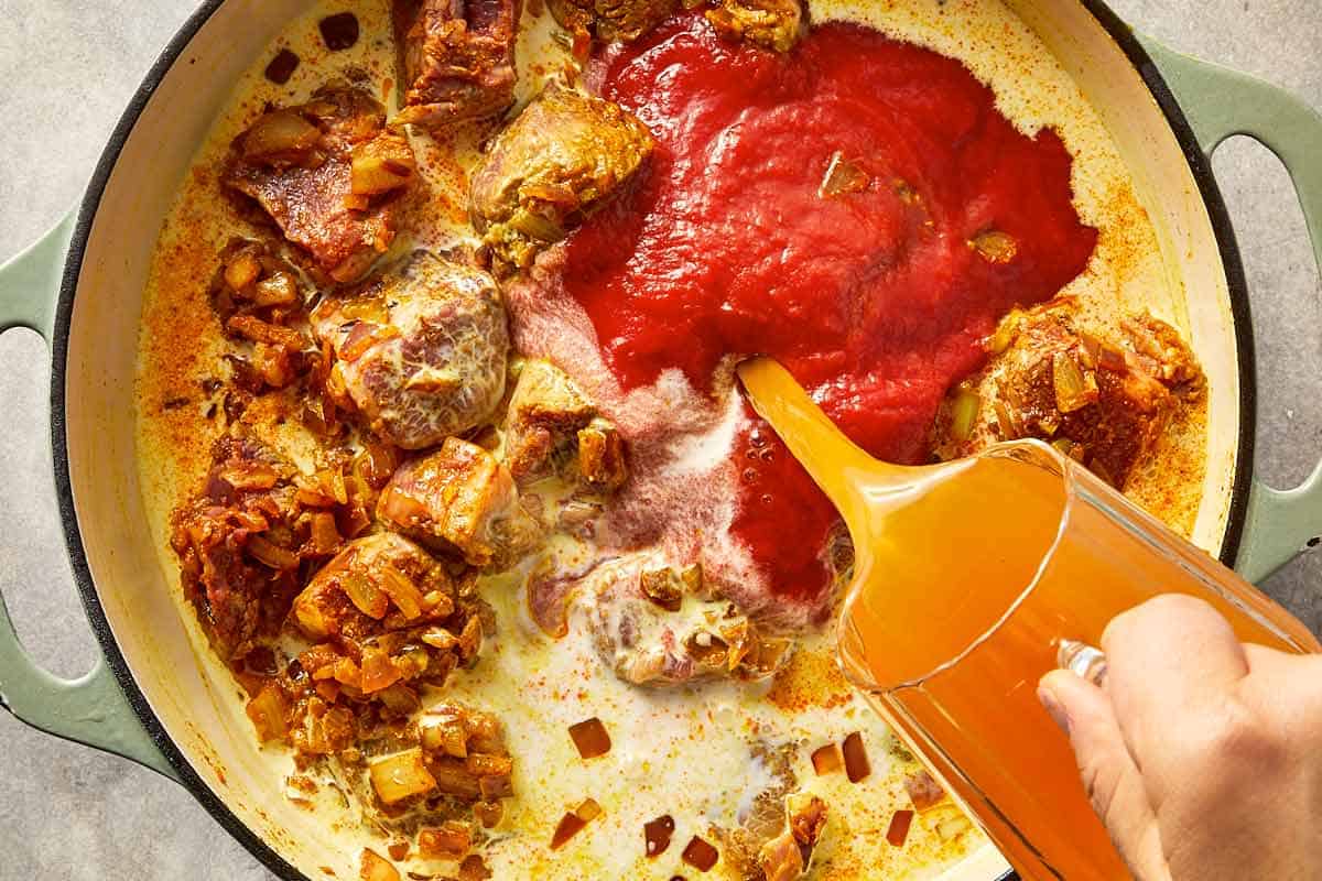 Tomato passata, coconut milk, and stock being poured into the pan with lamb.