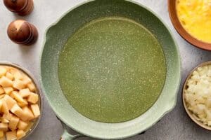 Olive oil heating in a nonstick pan for making Spanish Omelette with chopped potatoes and onion nearby.