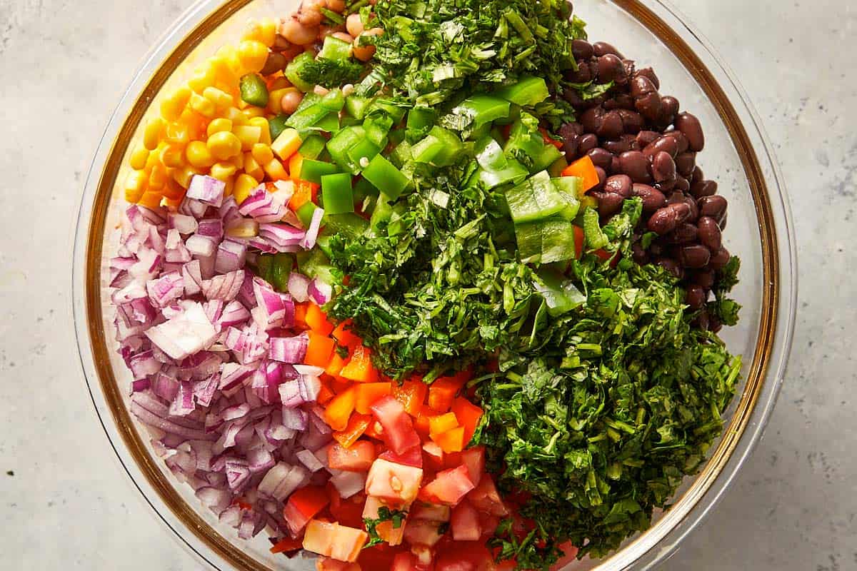 Black eyed pea salad ingredients in a bowl with black beans, corn, red onion, tomatoes, bell peppers, jalapeños and fresh coriander before mixing.