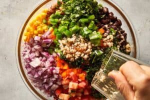 Black eyed pea salad ingredients with homemade lime vinaigrette being poured over beans, corn, tomatoes and fresh vegetables in a glass bowl.