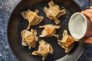 Pouring water from a small white cup into a hot skillet with dumplings to create steam.