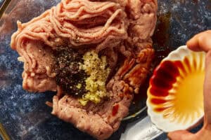 Pouring soy sauce into a bowl of raw ground turkey topped with garlic, ginger, and black bean paste.