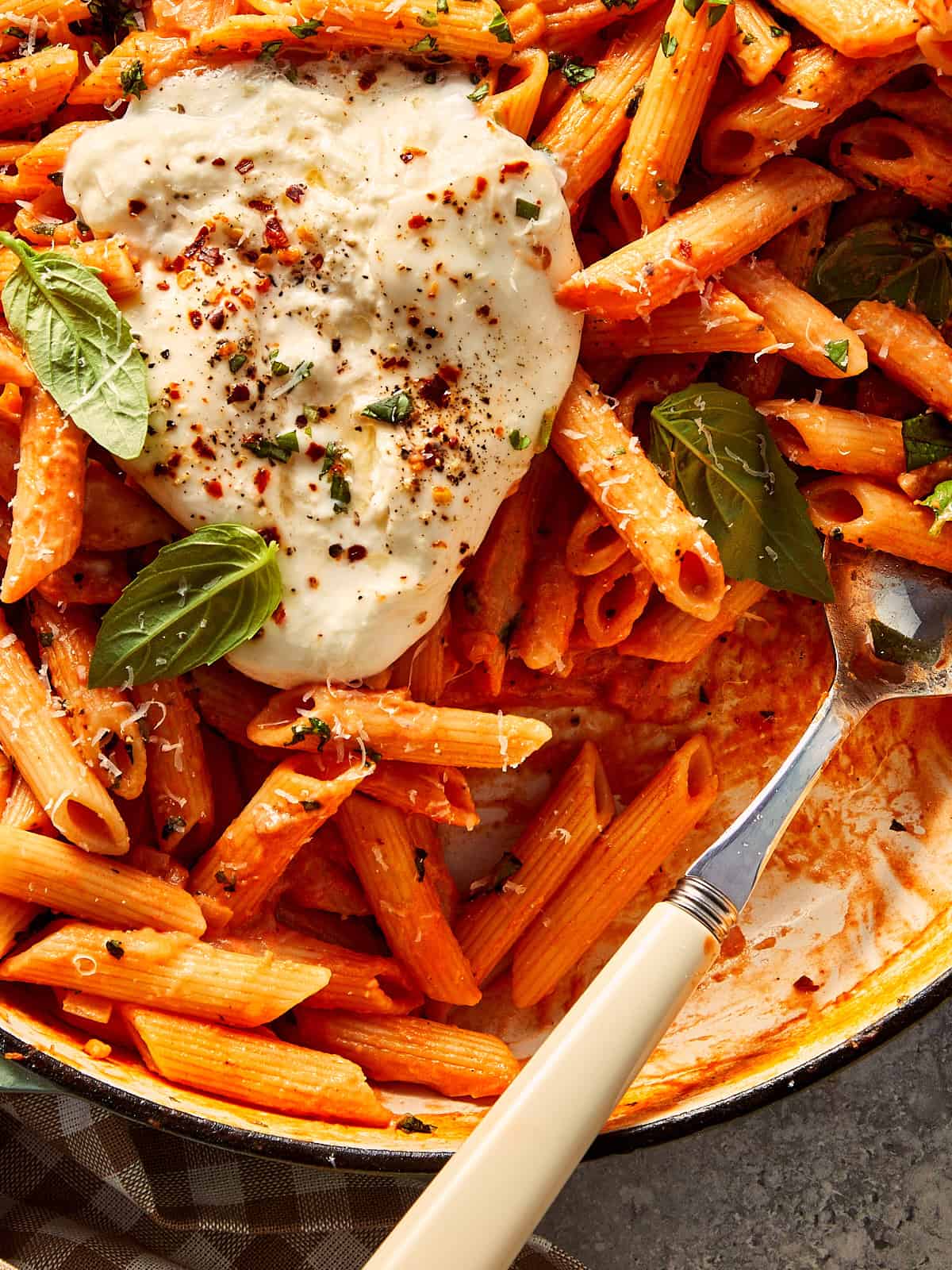Close-up of one-pot spicy vodka penne pasta in a pan, topped with a broken fresh burrata, fresh basil leaves, and chili flakes, with a serving spoon resting inside the dish.