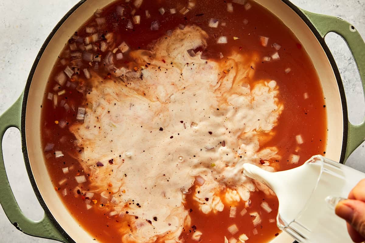 Pouring light cream into a green pot with tomato broth, shallots, and chili flakes to create the spicy vodka sauce base.